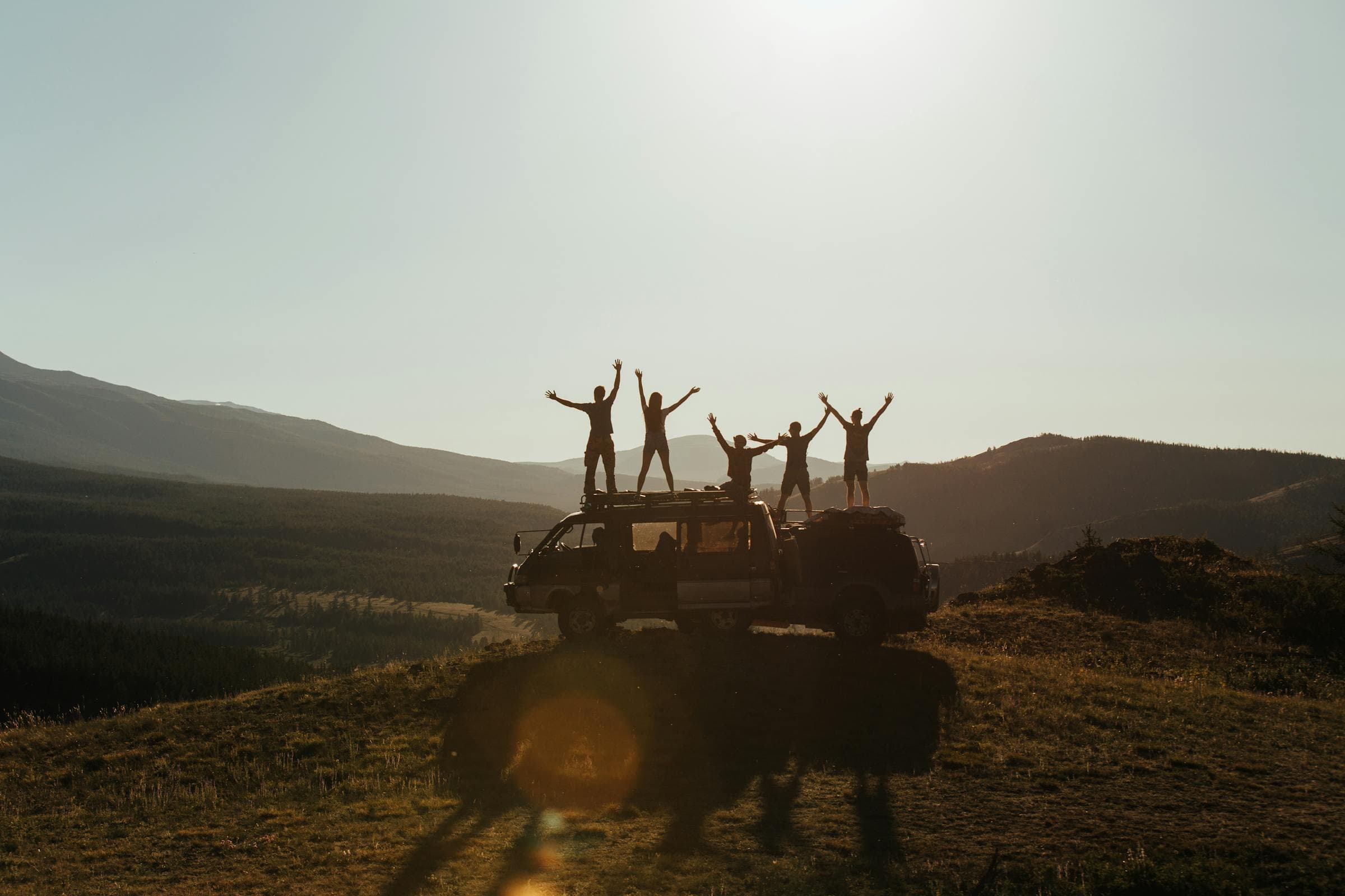 Silhouettes of a group standing on a van roof at sunrise, overlooking a mountain horizon: freedom, adventure, the next stage together